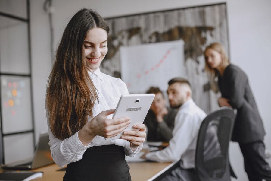Perempuan tersenyum membaca tablet, rekan timnya berdiskusi di belakang
