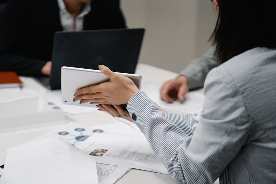 Tangan memegang tablet di meja rapat, rekan kerja membuka laptop di seberang